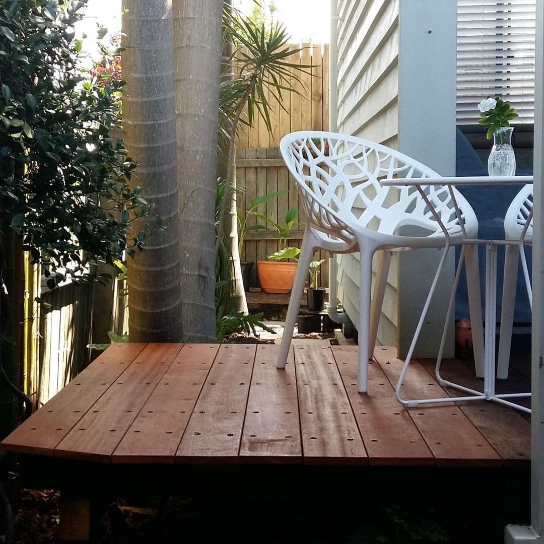 A wooden patio space in a small yard with a white bistro dining set surrounded by bamboo and palm trees.