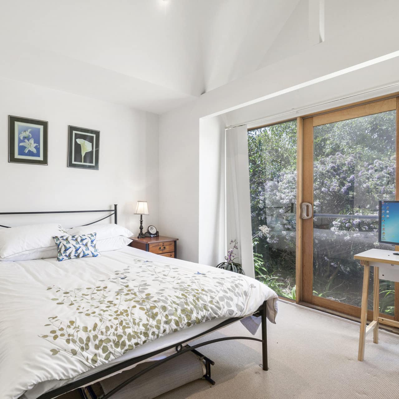 Sunny bedroom with a metal-framed king-size bed, wooden bedside table, desk and computer, tall windows, floral art.