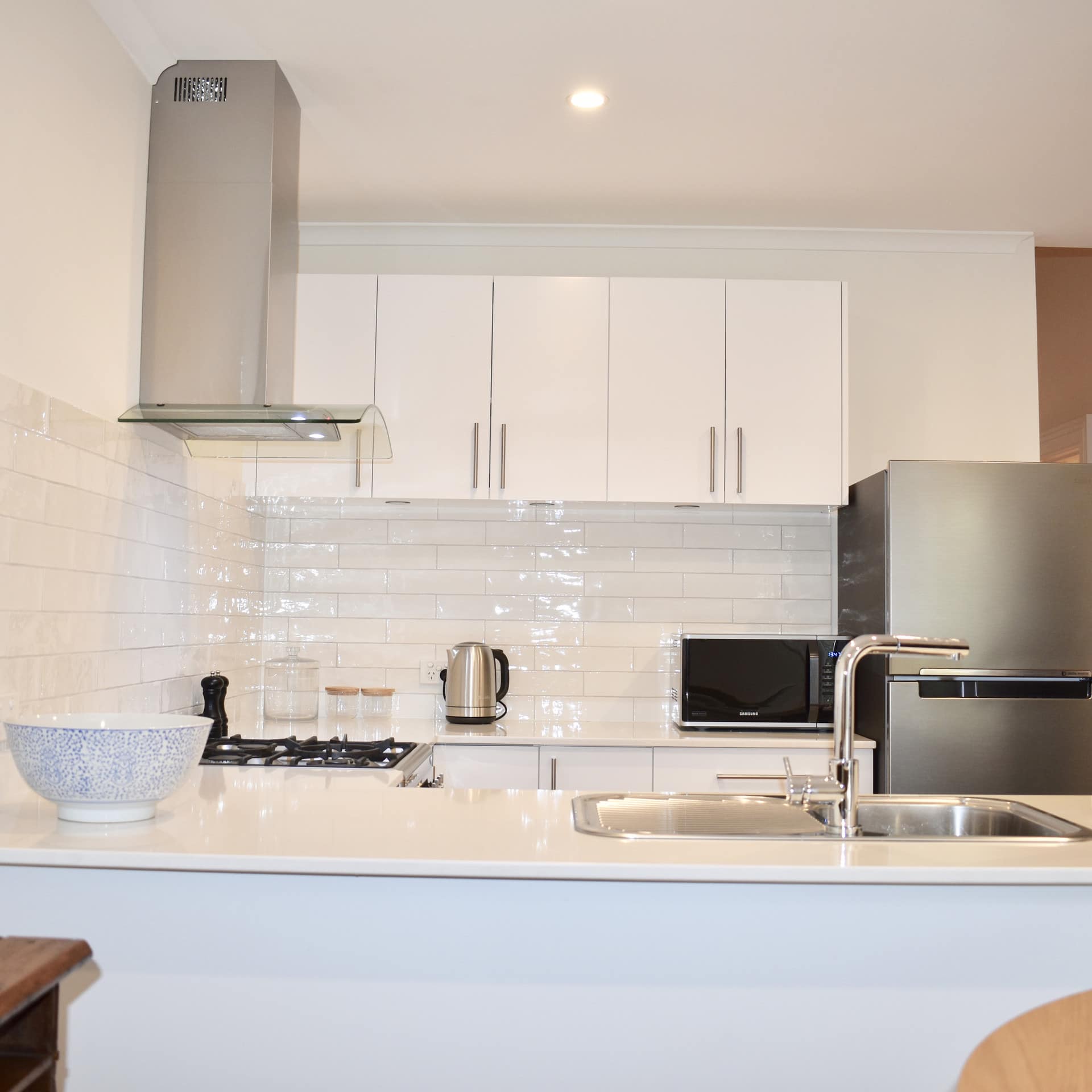 Stark white fully-equipped kitchen with shiny white brick splash guard wall and stainless refrigerator