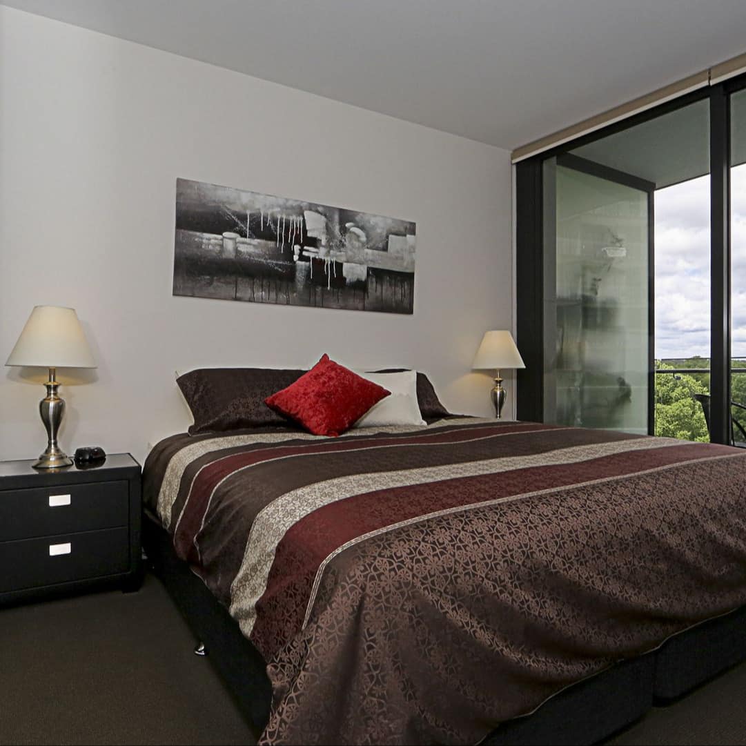 Neatly made king-size bed in front of floor to ceiling window that opens to a patio overlooking lush green trees