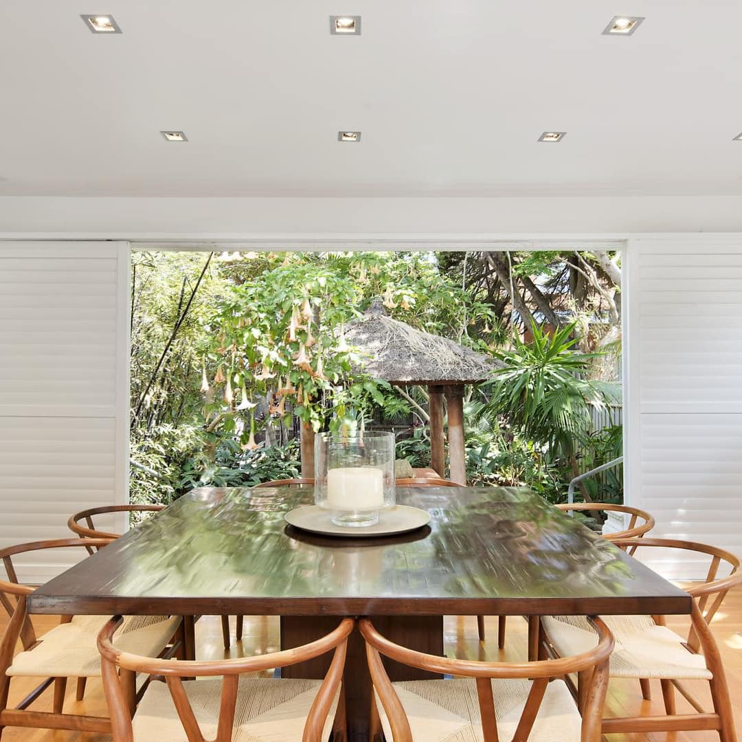 Sunny dining room with hardwood floors, wooden dining set, and open sliding doors to lush backyard.