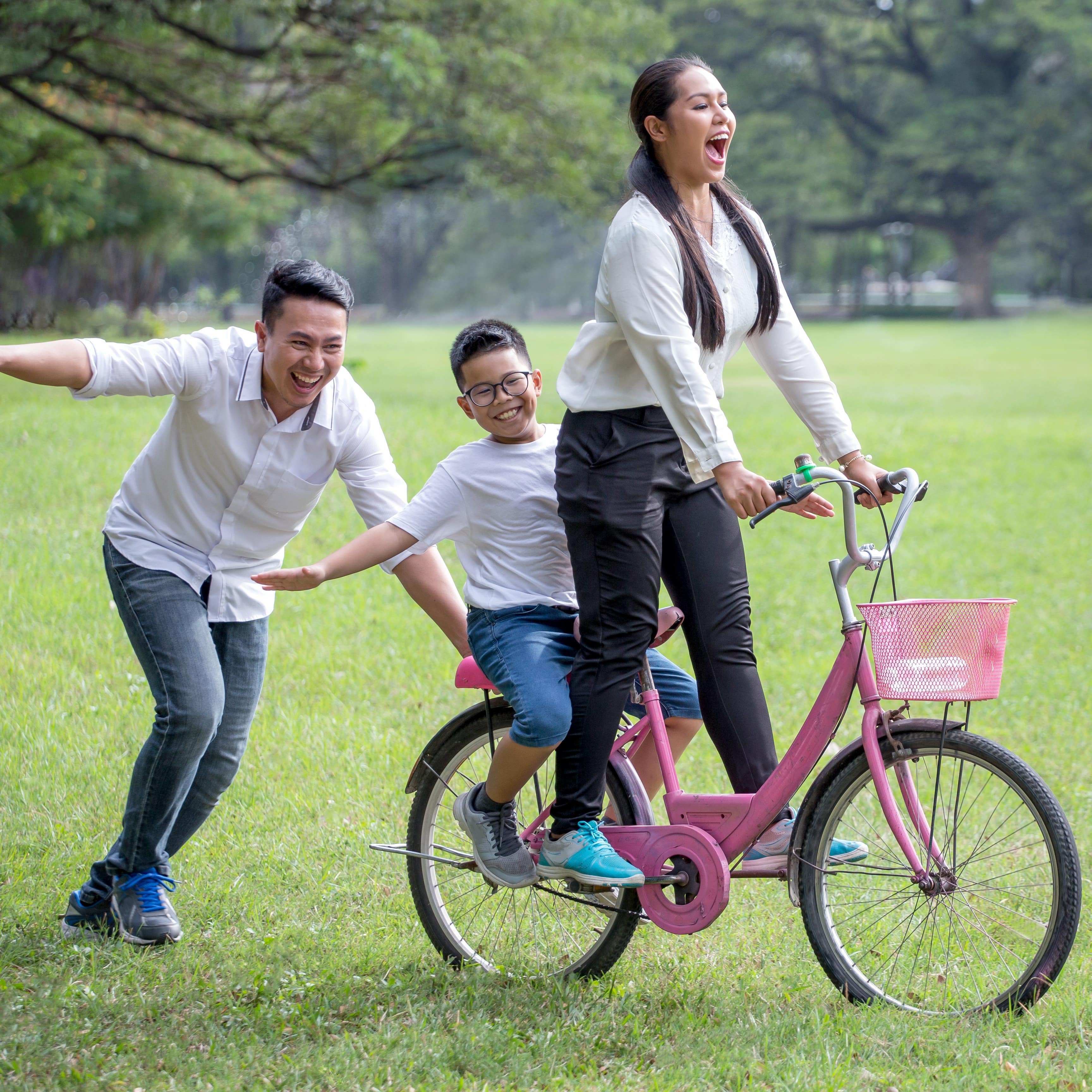 A father pushes mother and son on pink bicycle having fun and laughing in park
