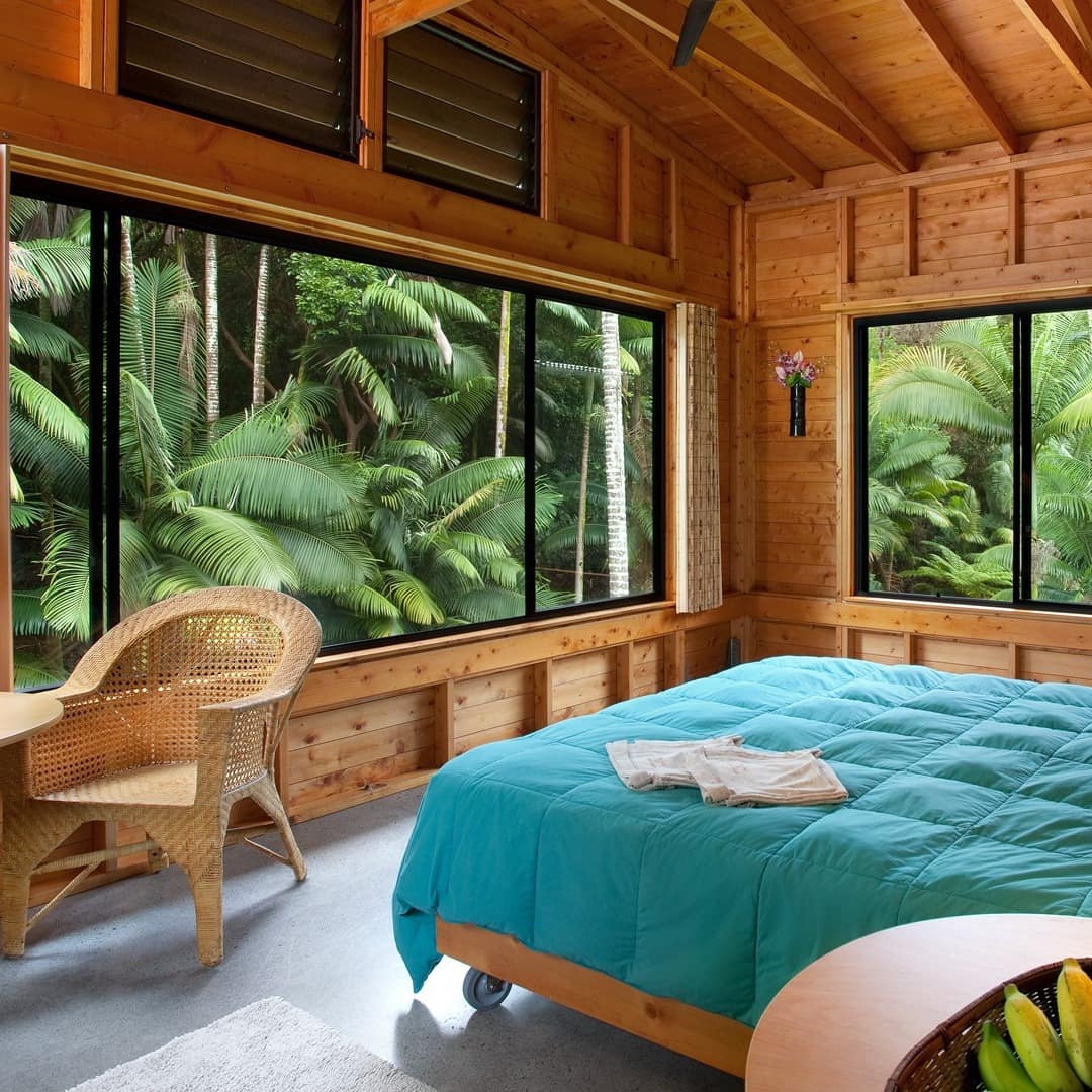 Light wood panelled bedroom with blue duvet bedcover and 2 large windows open to palm trees fronds filling the view.