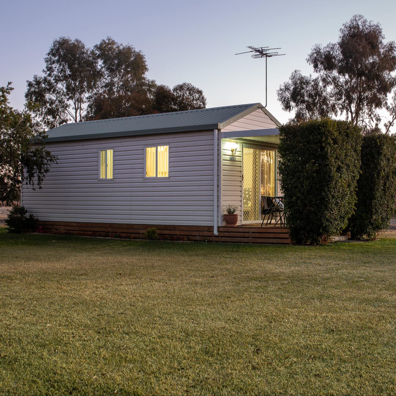 Exterior view of a cottage with white vinyl siding, green roof, covered patio space secluded behind manicured bushes.