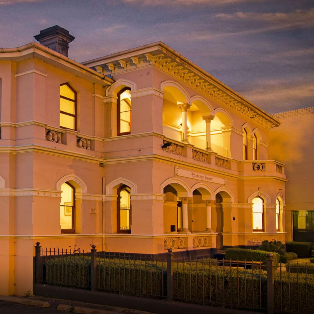 Nighttime view of a large Victorian apartment building with verandas on the second-story and ground level.