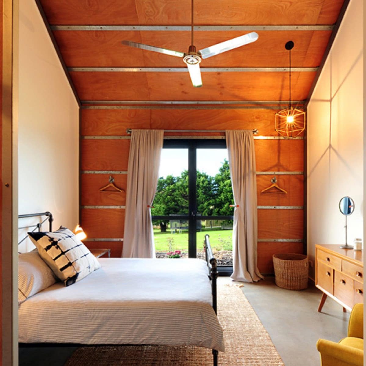Bedroom with a king size bed, modern furniture, wood panelled accent wall, grey stone floors, sliding glass doors.