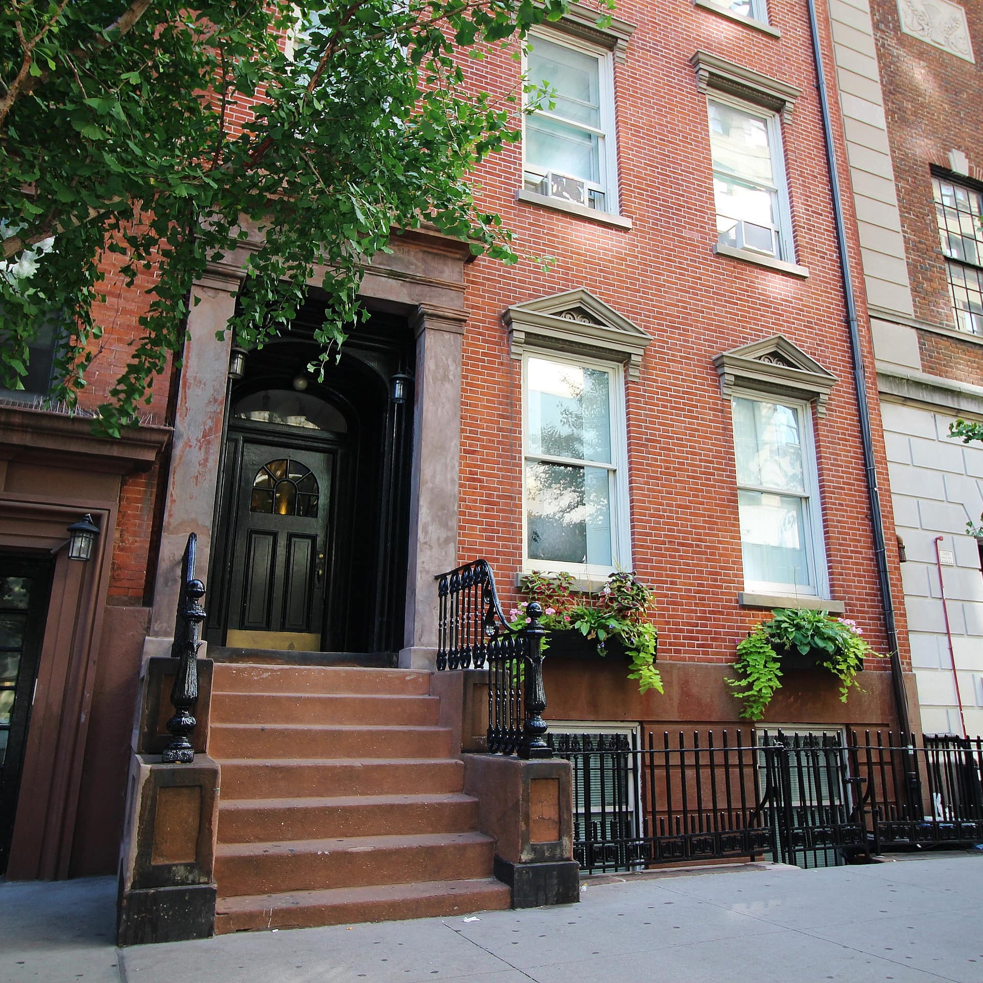 Brick townhouse with brown stairs to a black front door and overflowing window flower boxes.