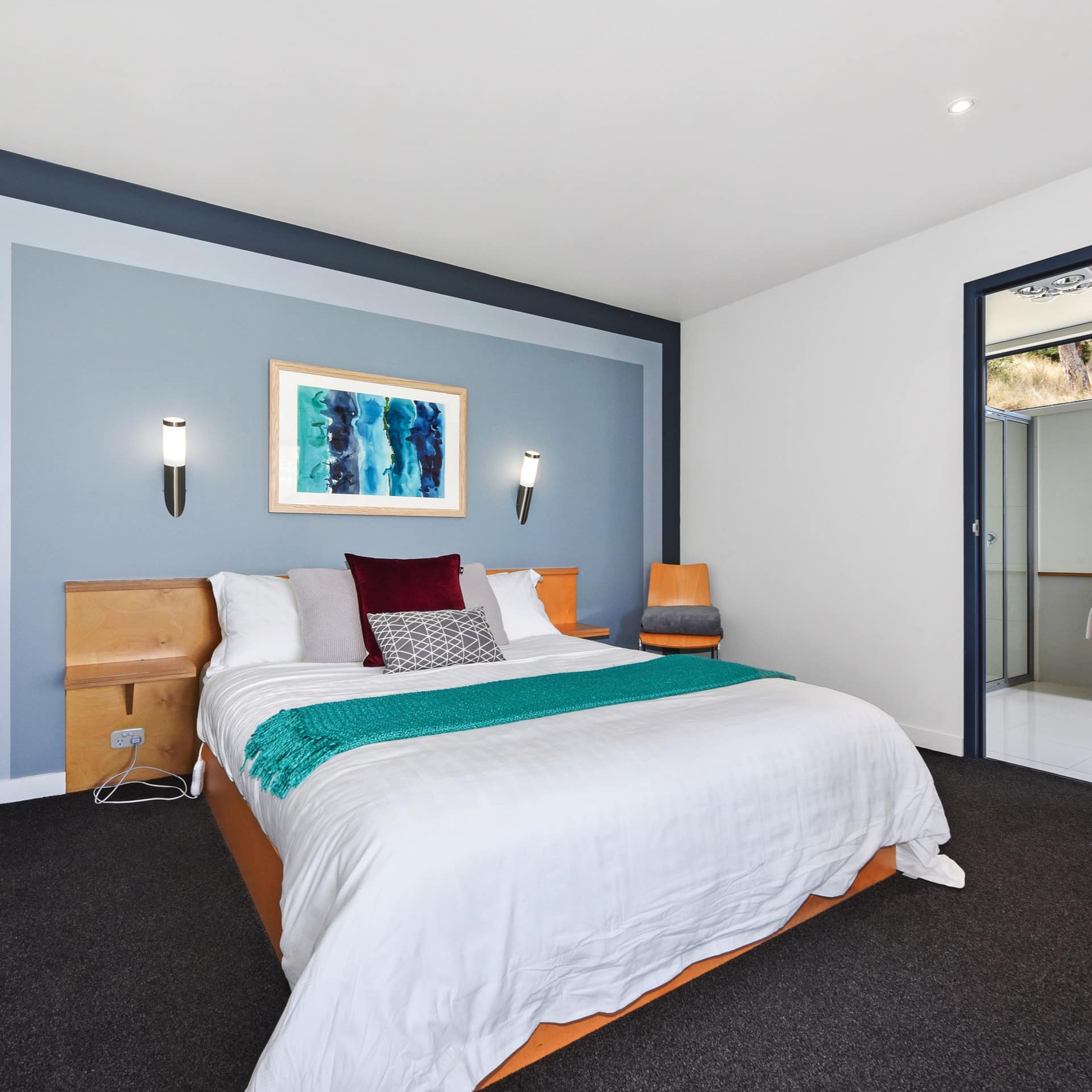 Modern bedroom with black carpet, blue accent wall, a king-size bed, white bedding, teal blanket, blue abstract painting.
