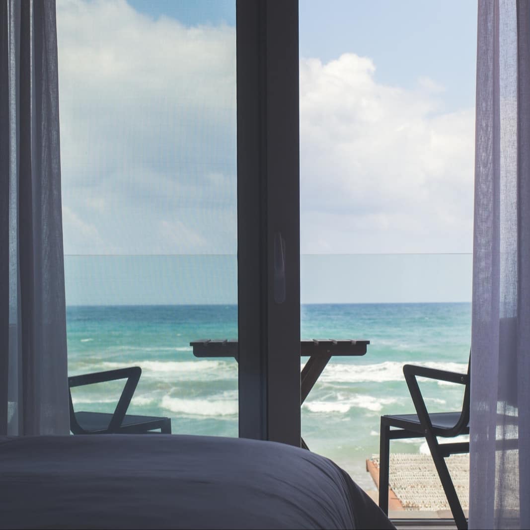 Bedroom of beachfront apartment with sheer white curtains and windows to a patio overlooking the ocean with table and chairs