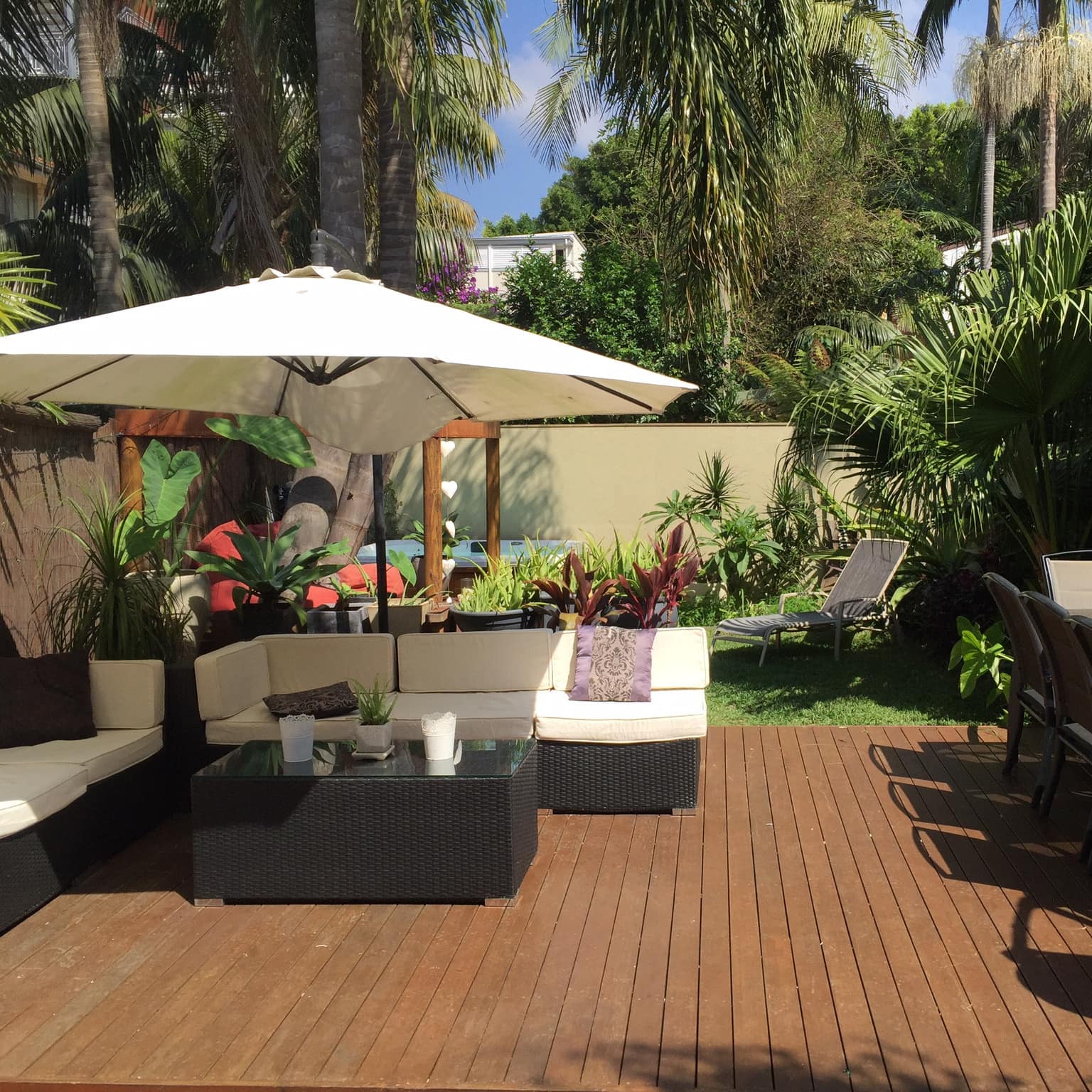 Private garden and deck with wicker outdoor sofas, shade umbrella and a dining table by a small green lawn with palms