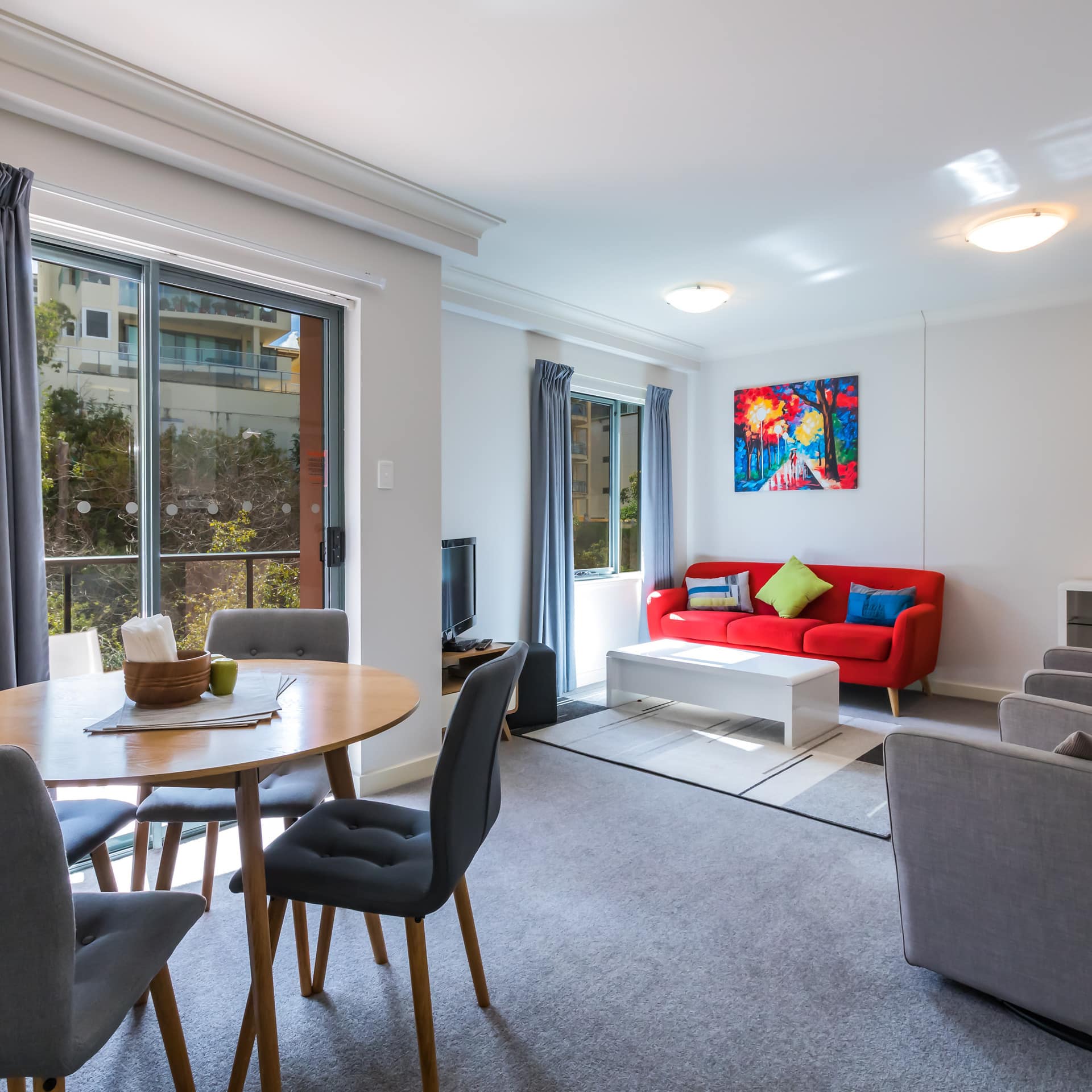 Open living and dining room with grey carpet, wooden dining set, red sofa and grey chairs, sliding glass doors to balcony.