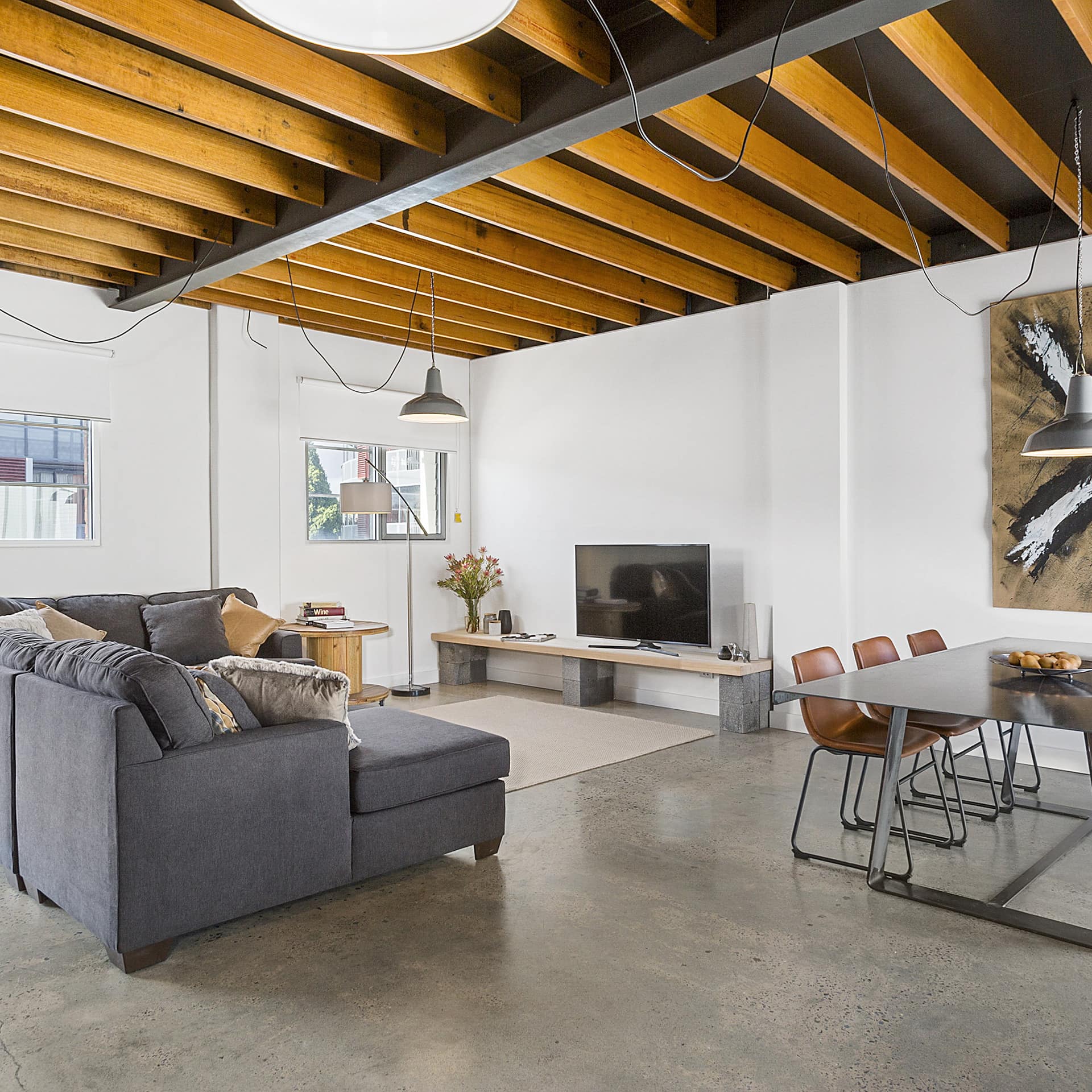 Modern living and dining room with grey cement floors, high ceilings with exposed beams, grey sectional, metal dining table.