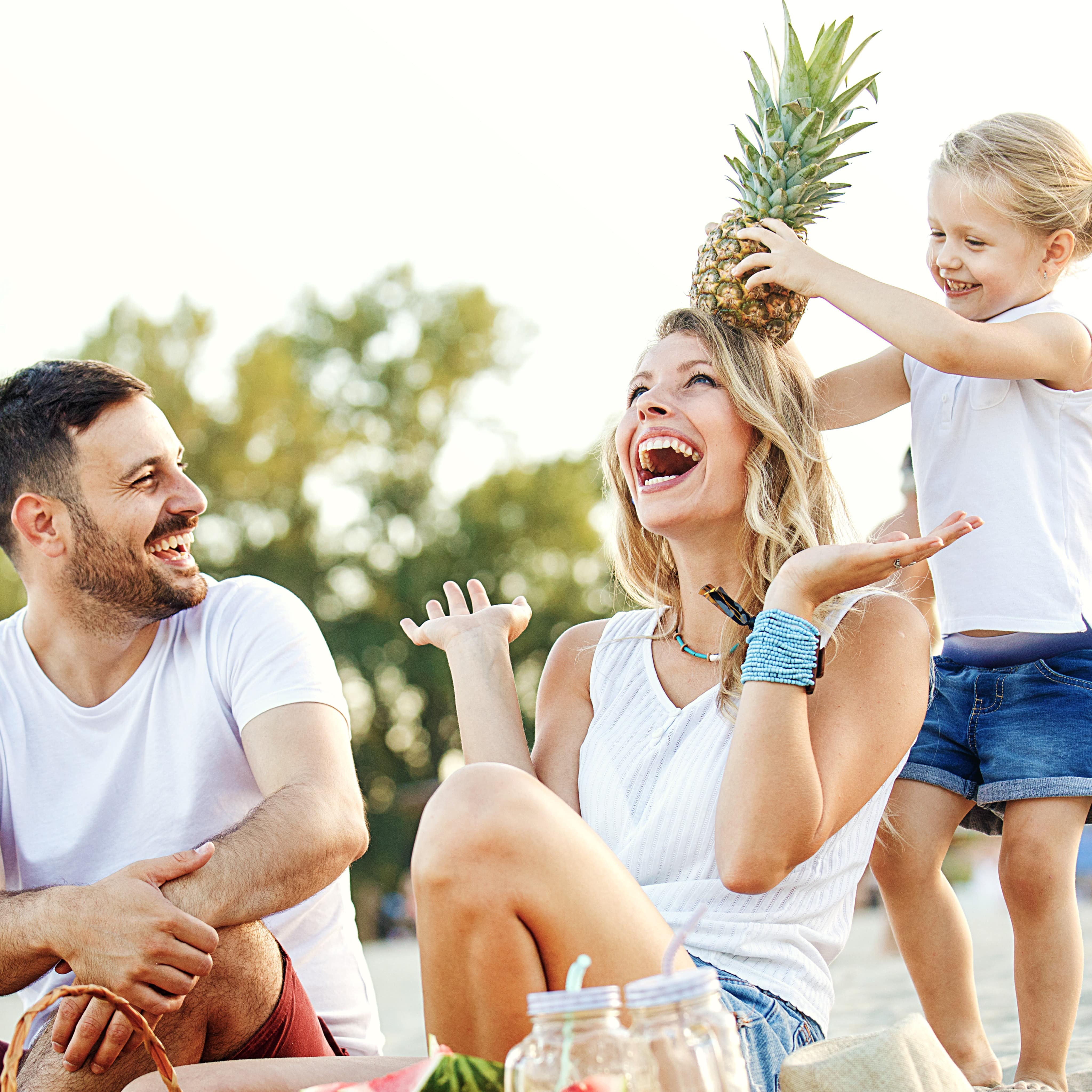 Family laughing on picnic while little girl puts a pineapple on her mom’s head