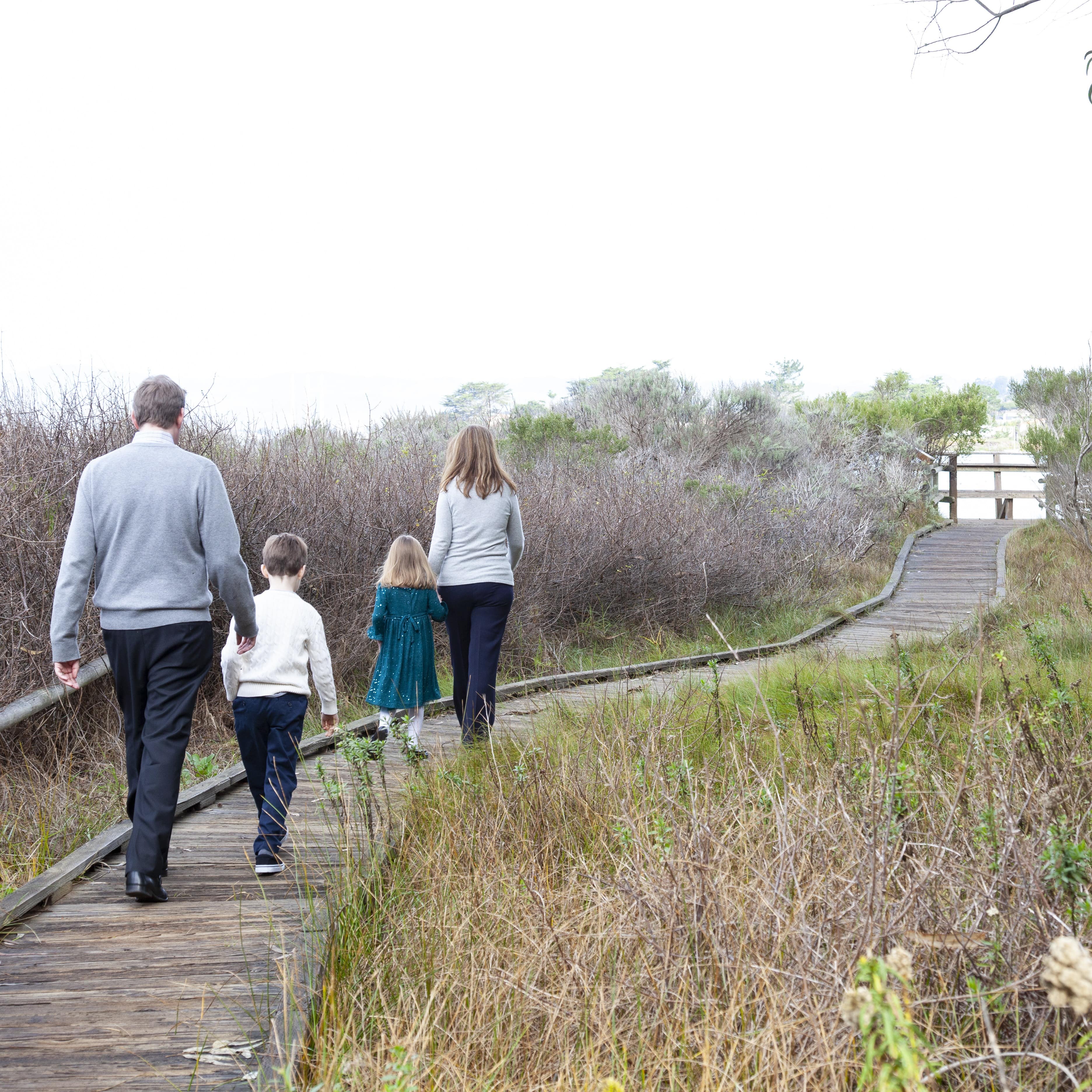 Family of four walking together away from viewer, on boardwalk through tall grasses in autumn