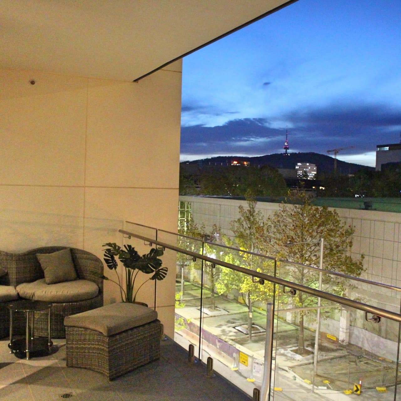 Nighttime view of a balcony with glass railings, wicker furniture, and city views.