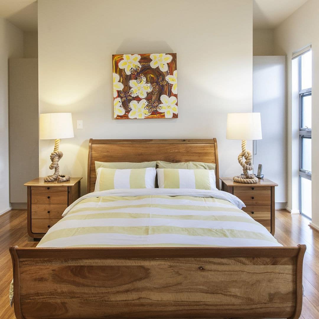 Sunny bedroom with hardwood floors, white walls, wooden furniture, king-size bed, yellow and white bedding.