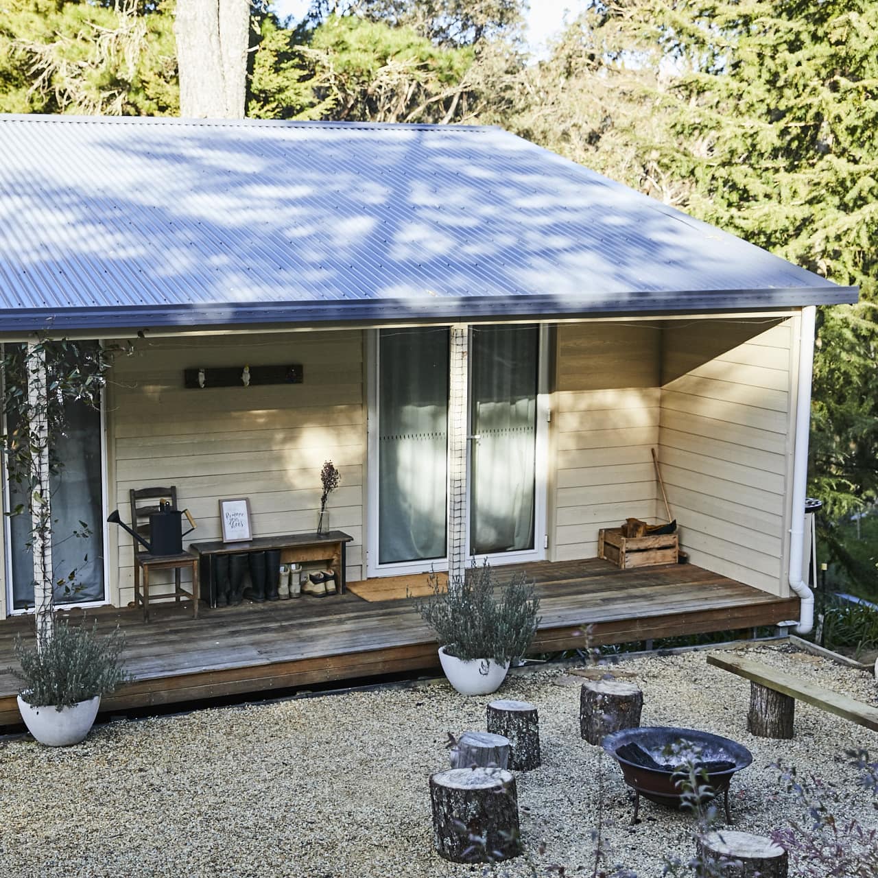 Rustic, Danish-style cabin with beige panel siding, tin roof, hardwood deck, pebble gravel and firepit with tree-stump seats.