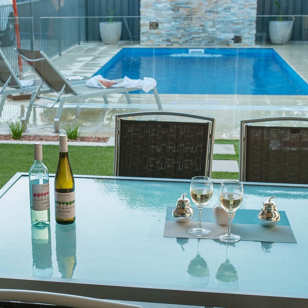 View of a glass dining table with wine glasses on a grey stone patio in front of a private pool.