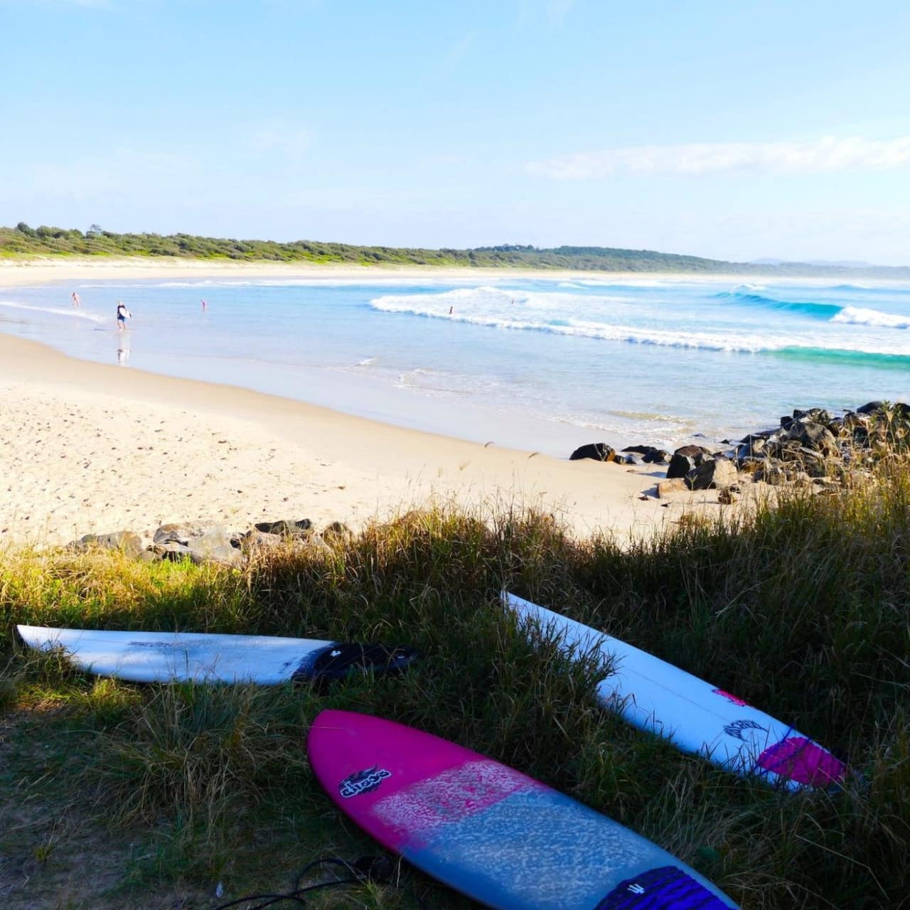 View of Crescent Head beach with crystal blue waves, a family walking in the sand, and surfboards lying in beachgrass.
