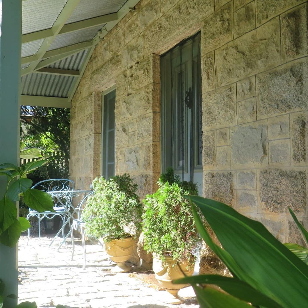View of a covered verandah with natural stone siding, teal metal bistro dining set, potted plants and lush greenery.