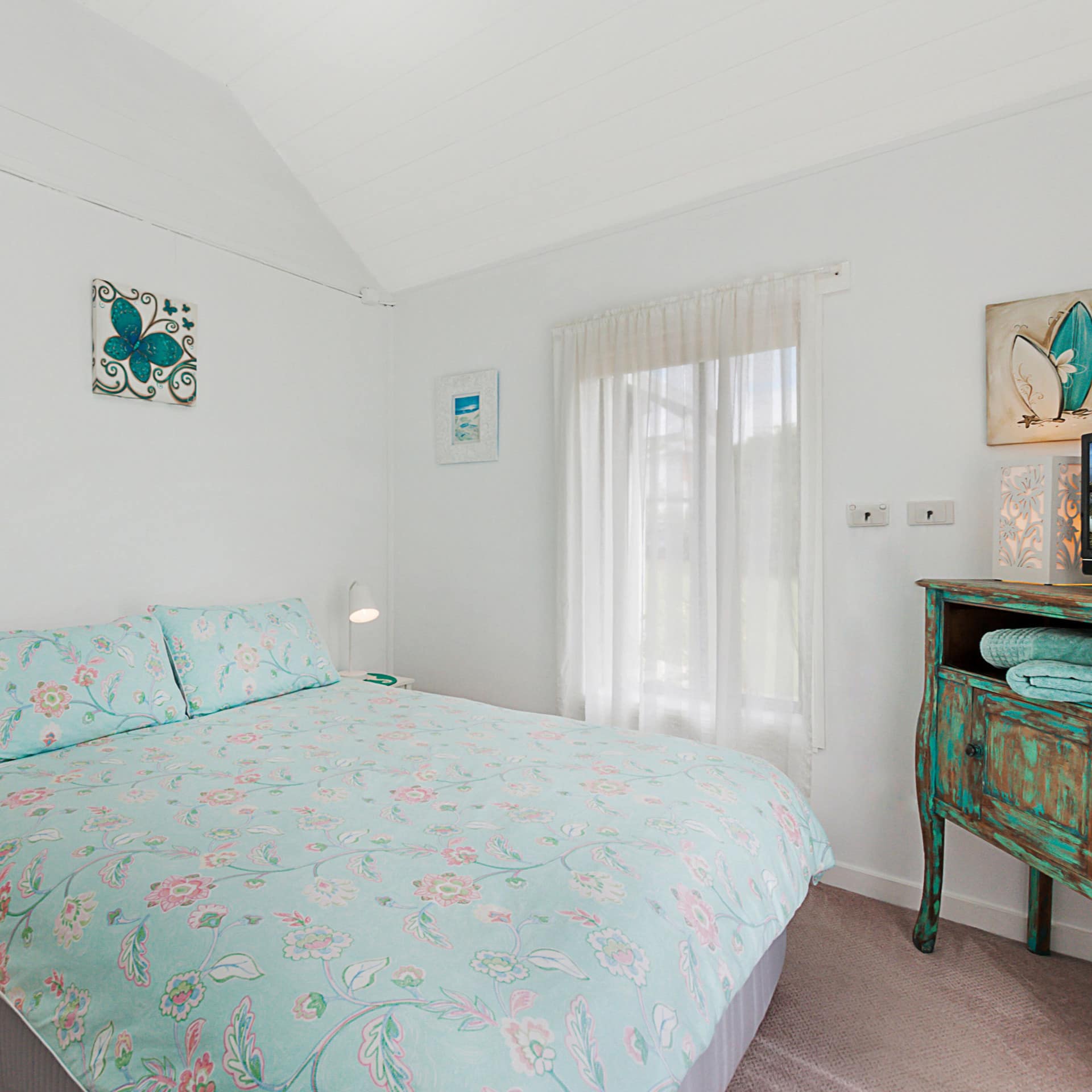 Bright bedroom, beige carpet, king size bed, teal floral bedding, reclaimed vintage vanity, tall window, sheer curtains.