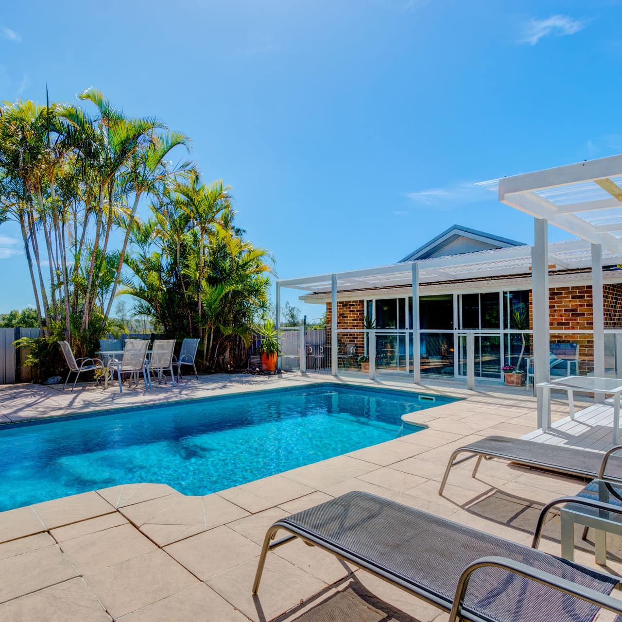 Fenced-in, back yard blue pool surrounded by patio furniture behind a small brick house with sliding glass doors