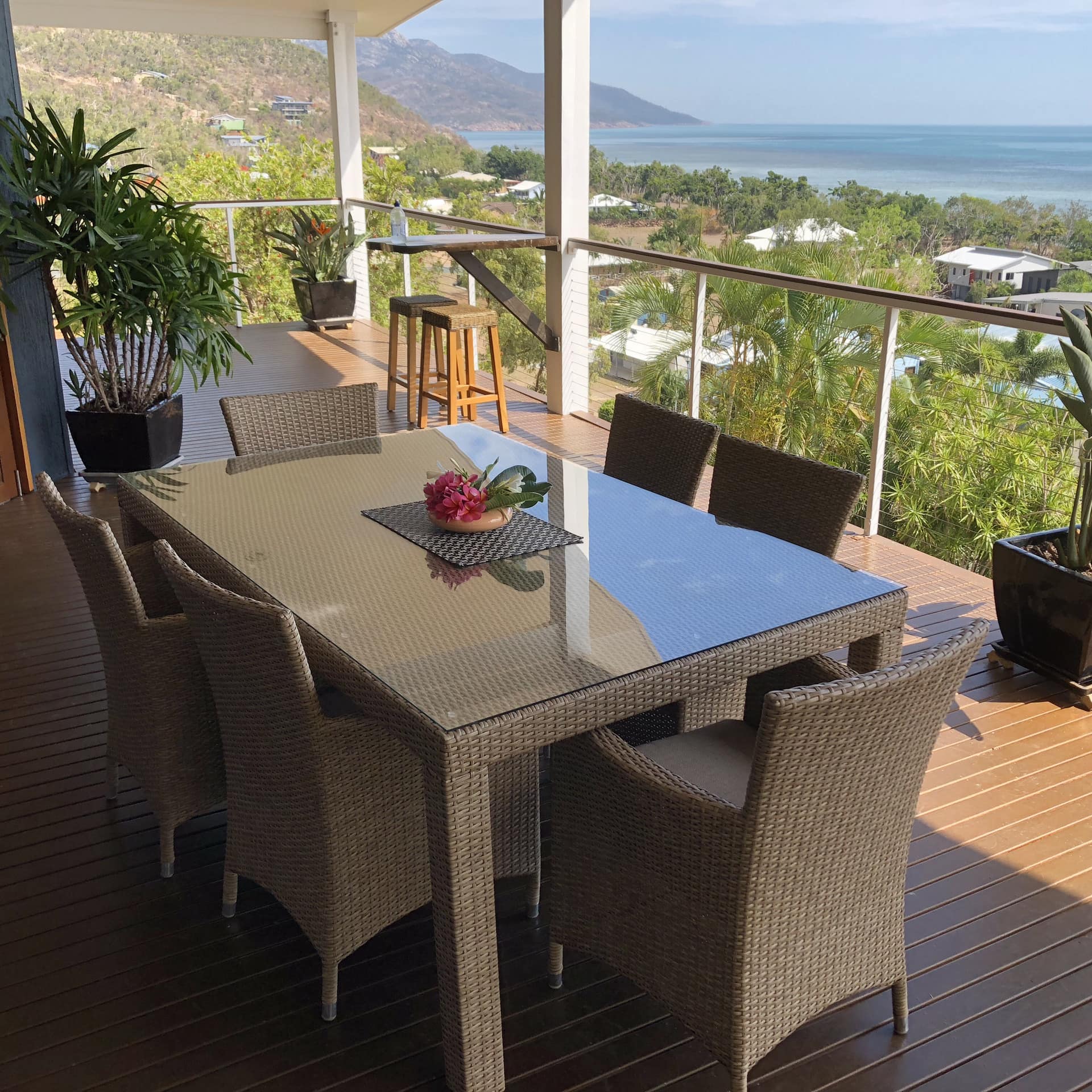 A covered veranda overlooking a neighborhood and the ocean, tan wicker dining set, wood floors, potted tropical plants.