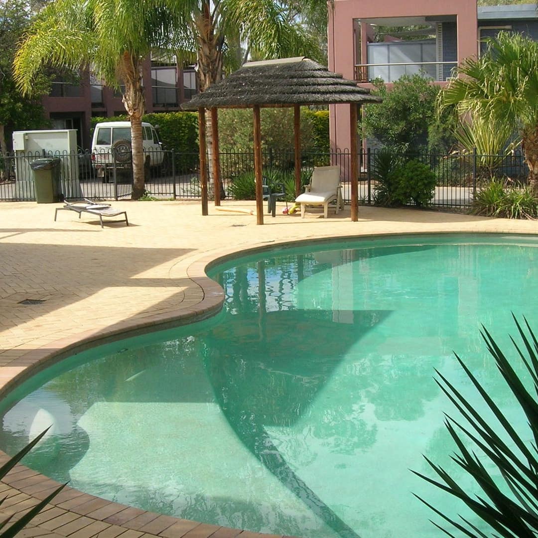 View of in-ground curved pool and beige tiled patio space decorated with tropical plants and enclosed by black iron fence.