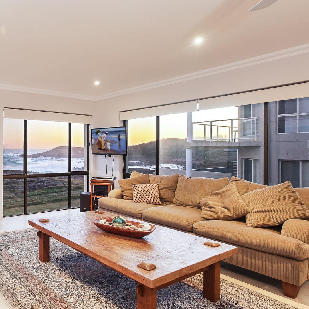 Spacious living room with tan plush sofa, wood coffee table and tall windows with panoramic views of the rocky ocean shore.