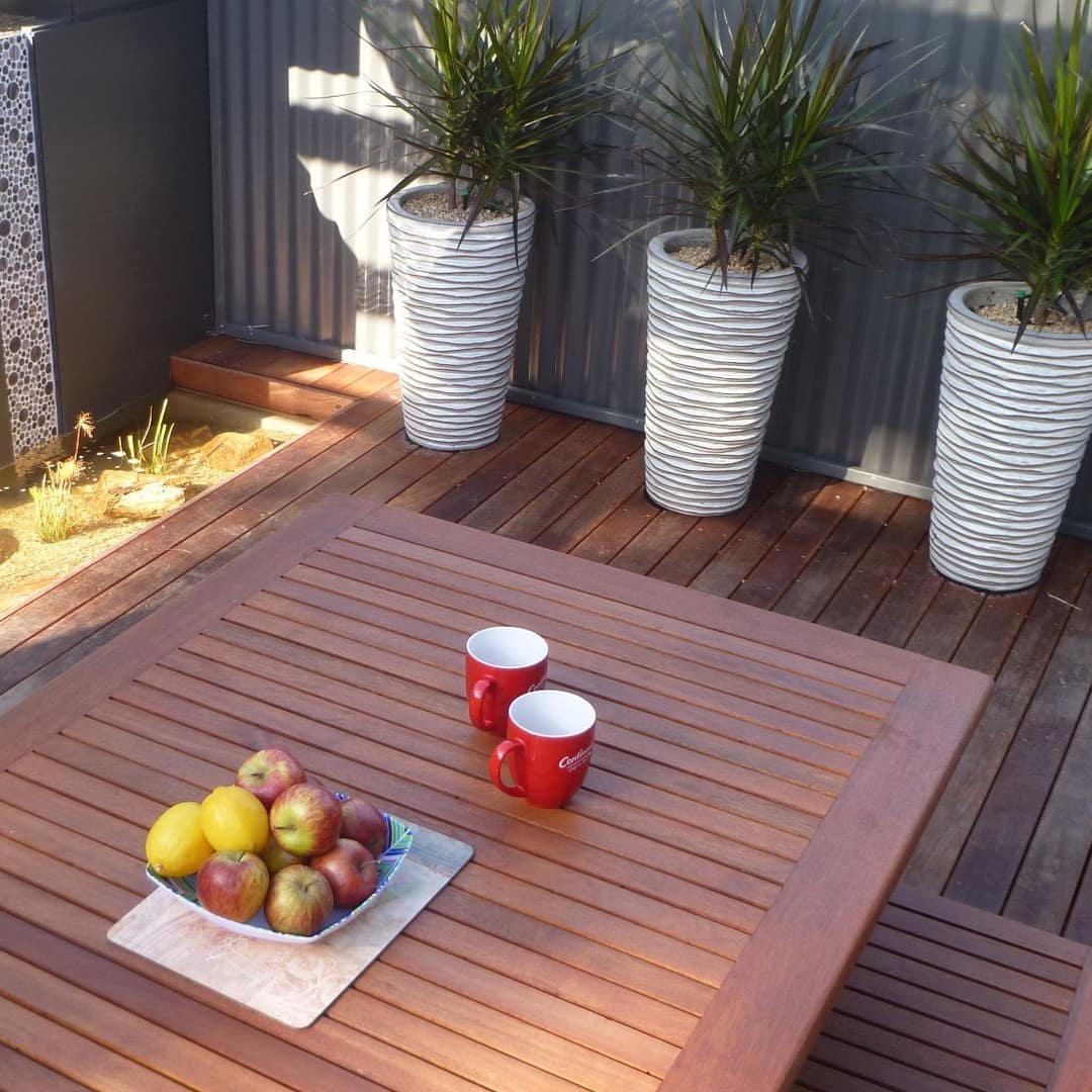 A wooden dining table with a bowl of apples and mugs on a wooden deck with a modern fountain and potted tropical plants.