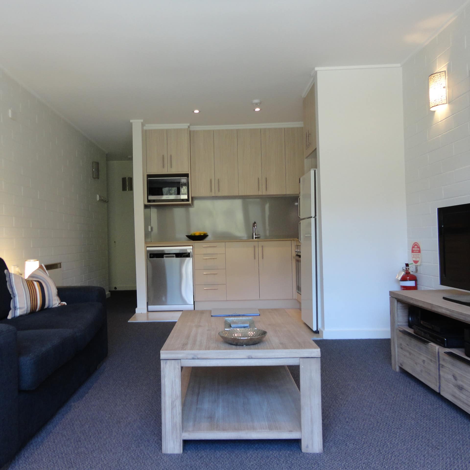 Open living room with grey carpeted floors, white brick walls, a kitchenette with wood cabinets, navy blue sofa.