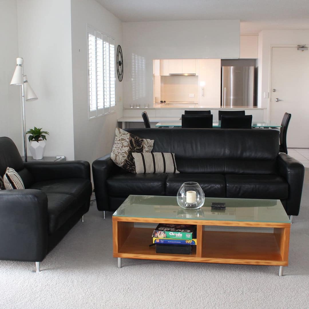Open living room and kitchen, white carpet and walls, black leather sofas, wood coffee table, windows, white ventian blinds.