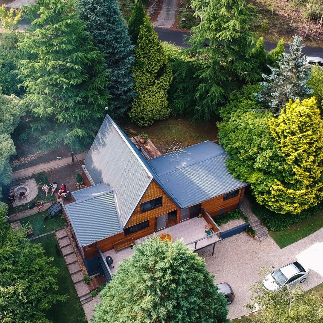 Aerial view of modern mountain home with A-frame roof and a secluded fire pit in the backyard surrounded by a grove of trees.