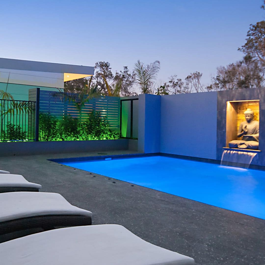 Night scene of a grey stone patio space, inground pool with buddha statue waterfall, white lounge chairs, tropical plants.