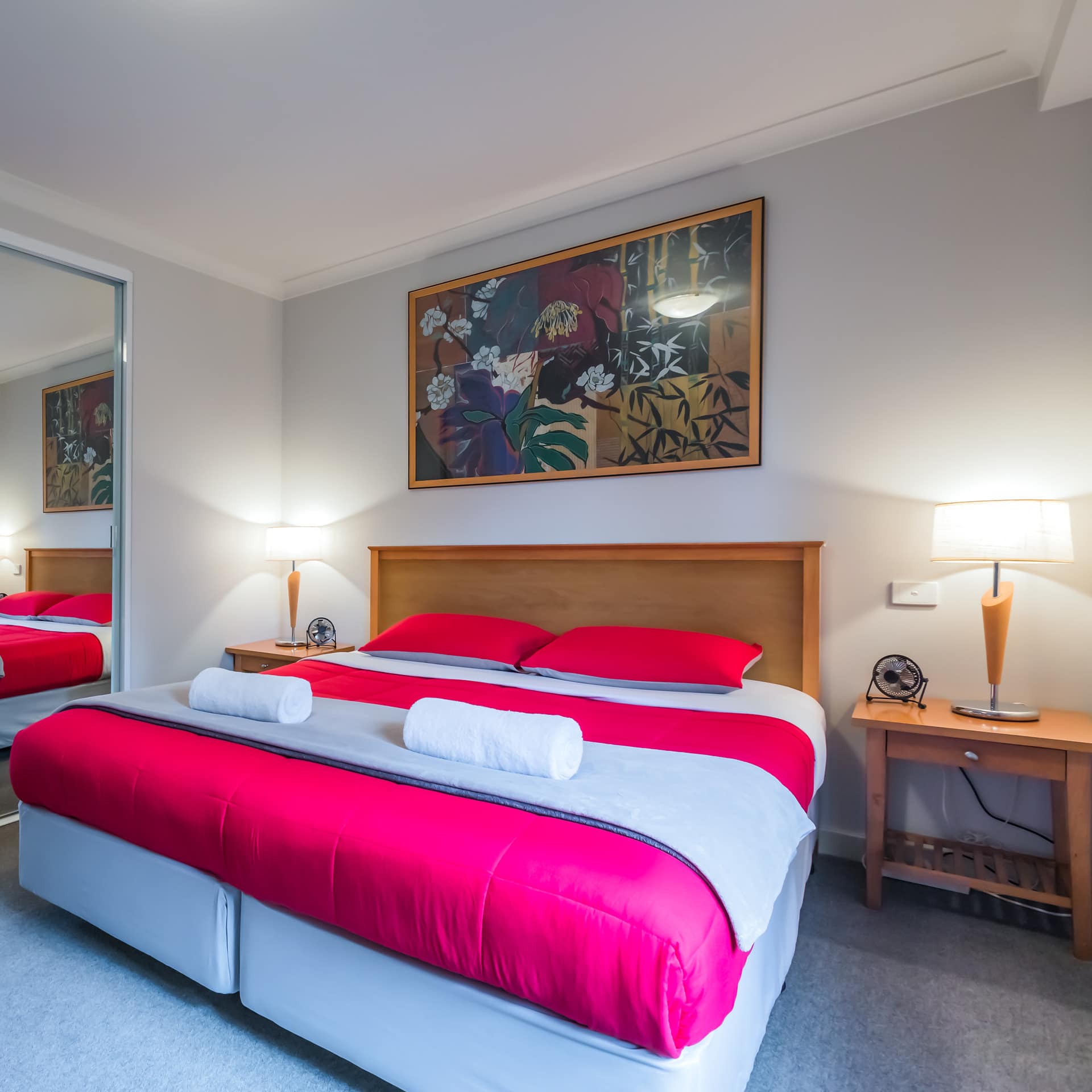 Bedroom with grey carpet, a king size bed, white and red bedding, floral painting, and mirrored closet doors.