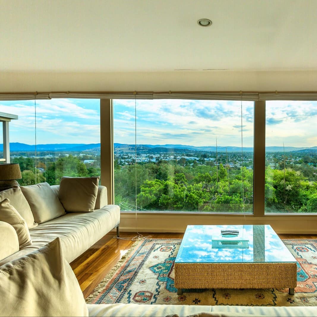Open living room plan with floor-to-ceiling windows, tan Scandinavian cloth sofas, a wicker table and a treetop view.