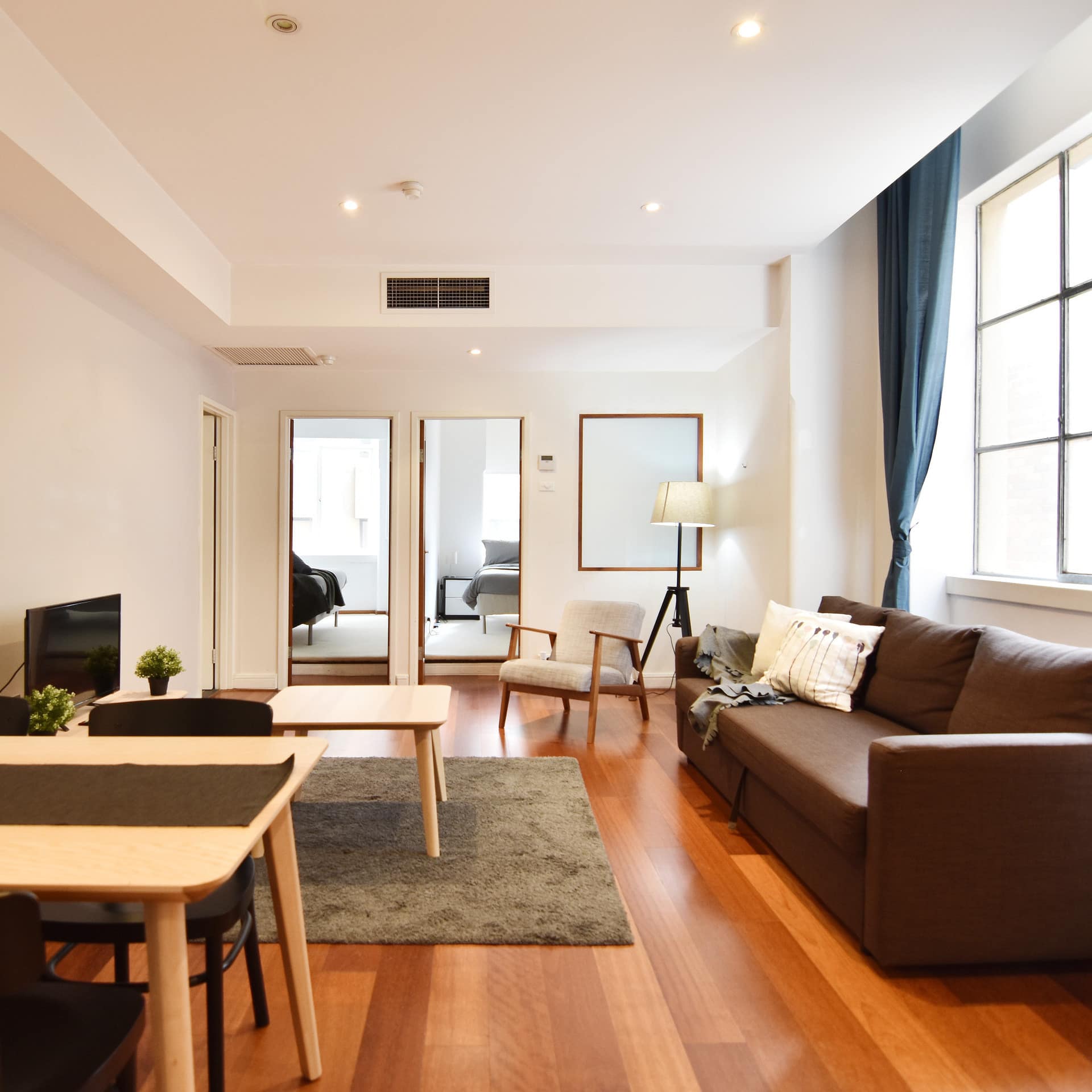 Open living room with brown sofa, wooden dining set, grey rug and wide windows with blue curtains.