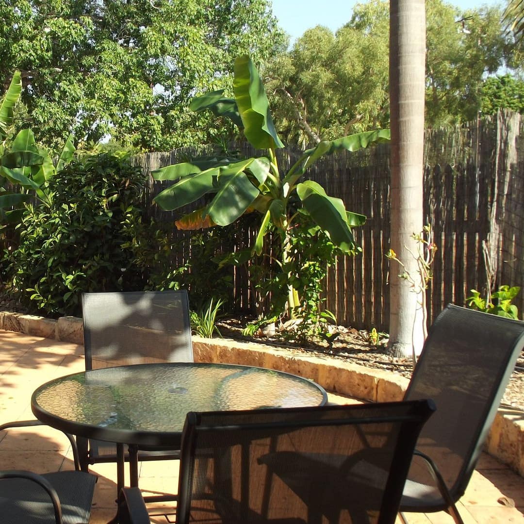 Sunny view of a patio space with tan tile, glass dining set and landscaped plant beds with tropical plants.