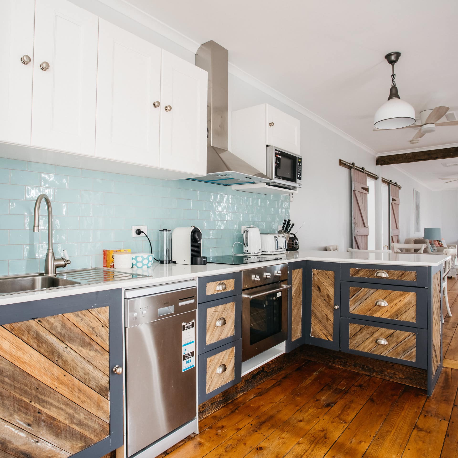 Open, gallery-style kitchen with hardwood floors, wood panelled cabinets, teal backsplash, and stainless steel appliances.