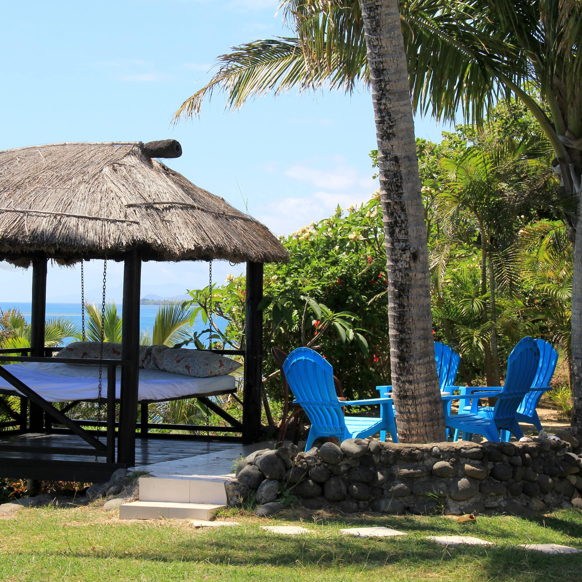 A straw hut cabana with a hanging bed and a blue dining set, surrounded by palm trees with ocean views.
