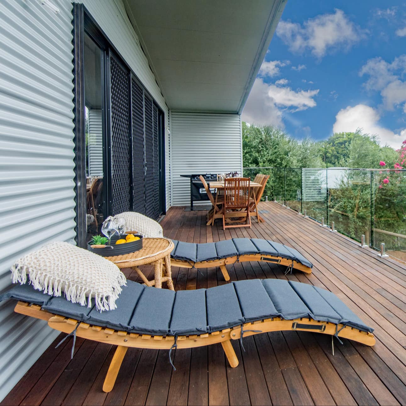 A wooden deck with glass railings, modern wooden lounge chairs, and wooden dining set, overlooking lush greenery.