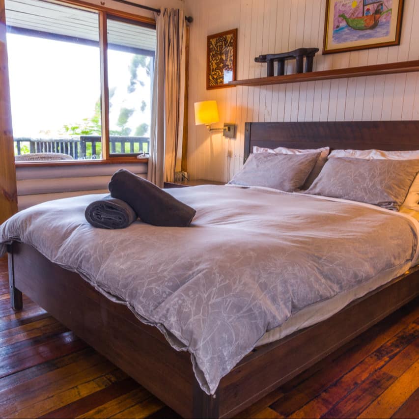 A rustic bedroom with hardwood floors, white panelled walls, a king size bed, grey bedding and tall windows.