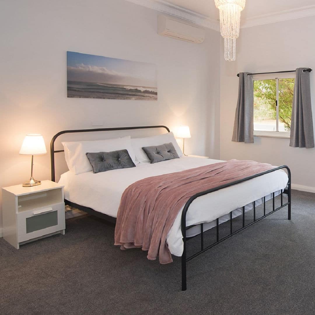 Spacious bedroom with grey carpet, white walls, a black metal framed king size bed, white and pink bedding, grey pillows.