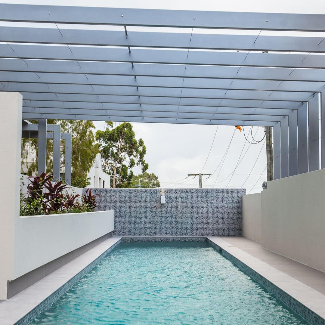 View of lap pool enclosed by white walls and metal beams, raised plant bed with purple plants, mosaic tile backsplash.