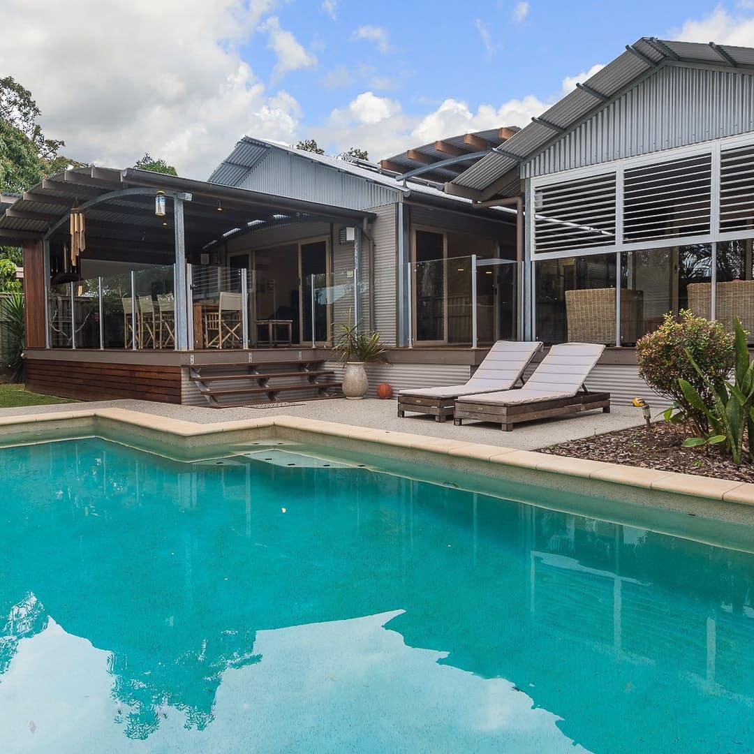 View of an inground pool with grey lounge chair on the pool deck next to a modern home.