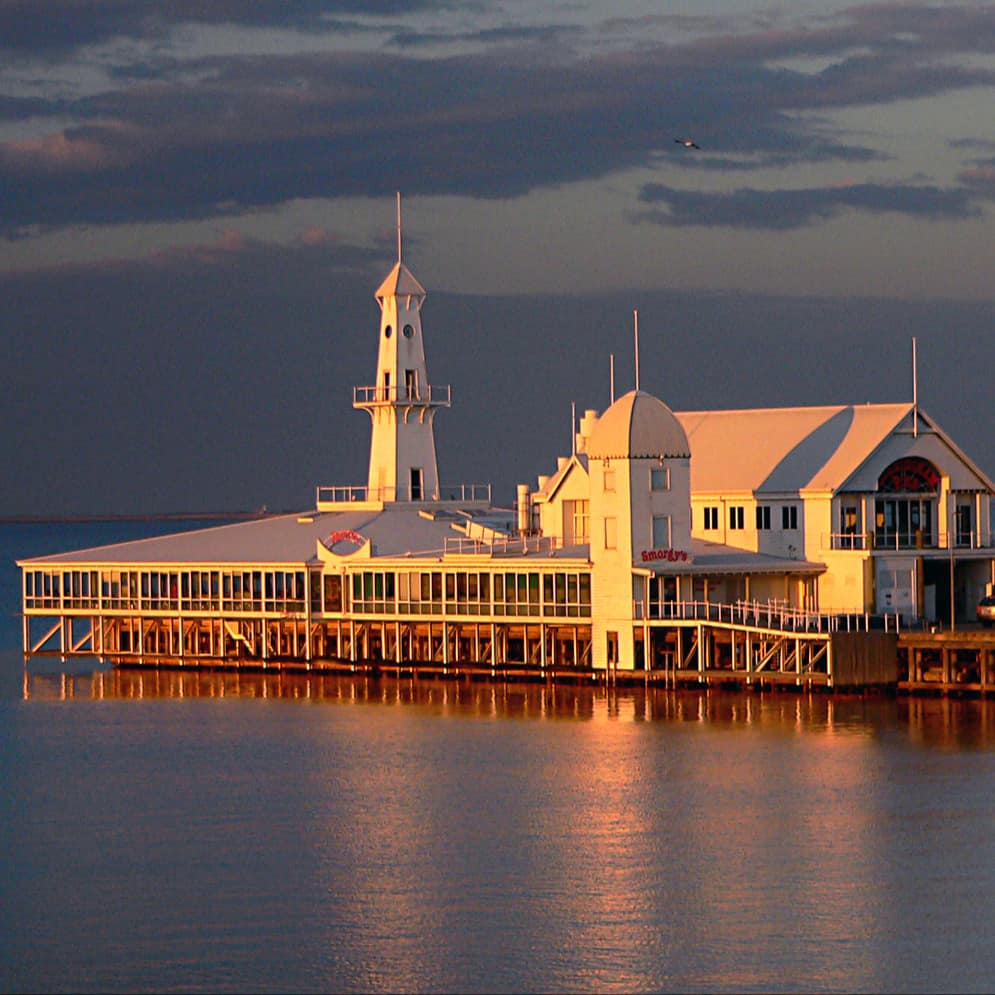 A view of Cunningham Pier in Geelong from the shore at sunset with a lighthouse and pavilion on the Corio Bay