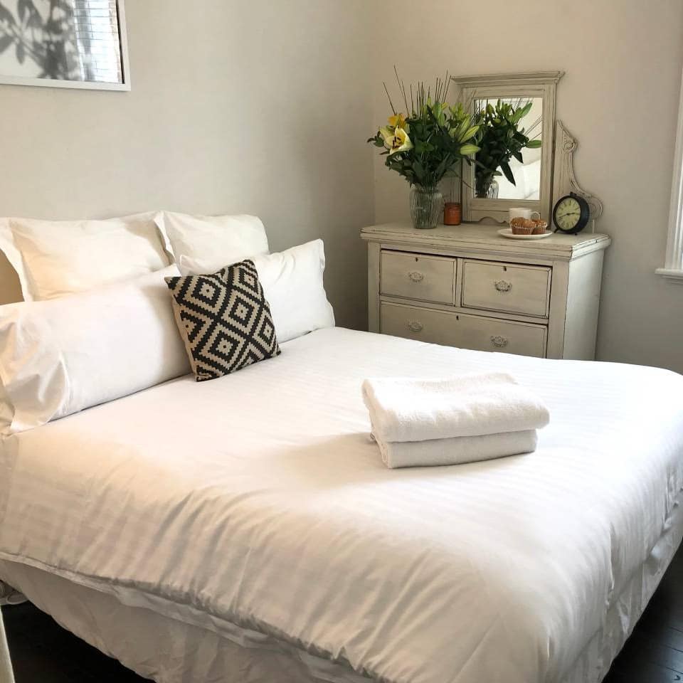 Bedroom with black hardwood floors, queen size bed, white linen, and a white vintage vanity with a bouquet of white lilies.