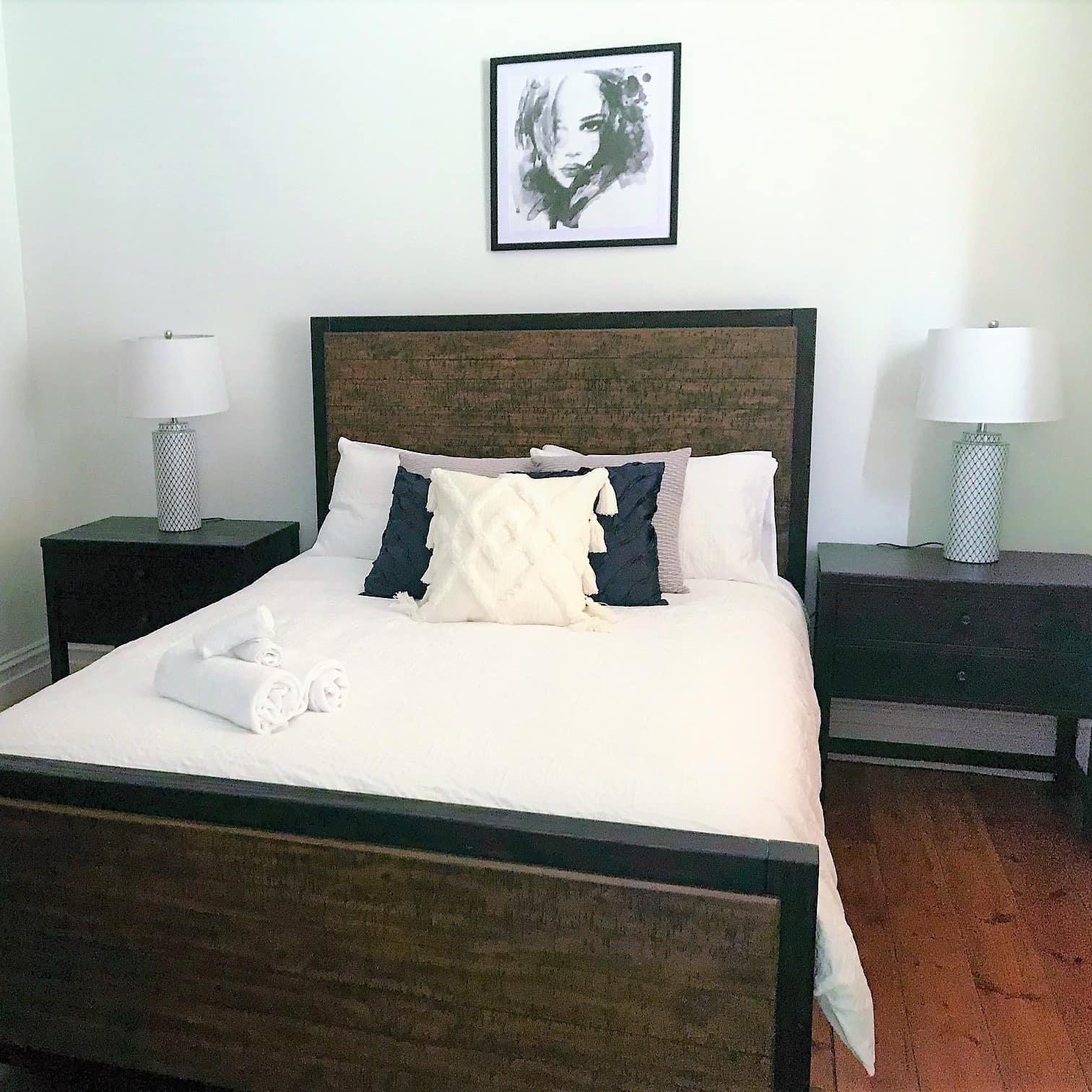 Bedroom with wood-framed queen size bed, white and grey bedding, black bedside tables, rolled towels on the bed.