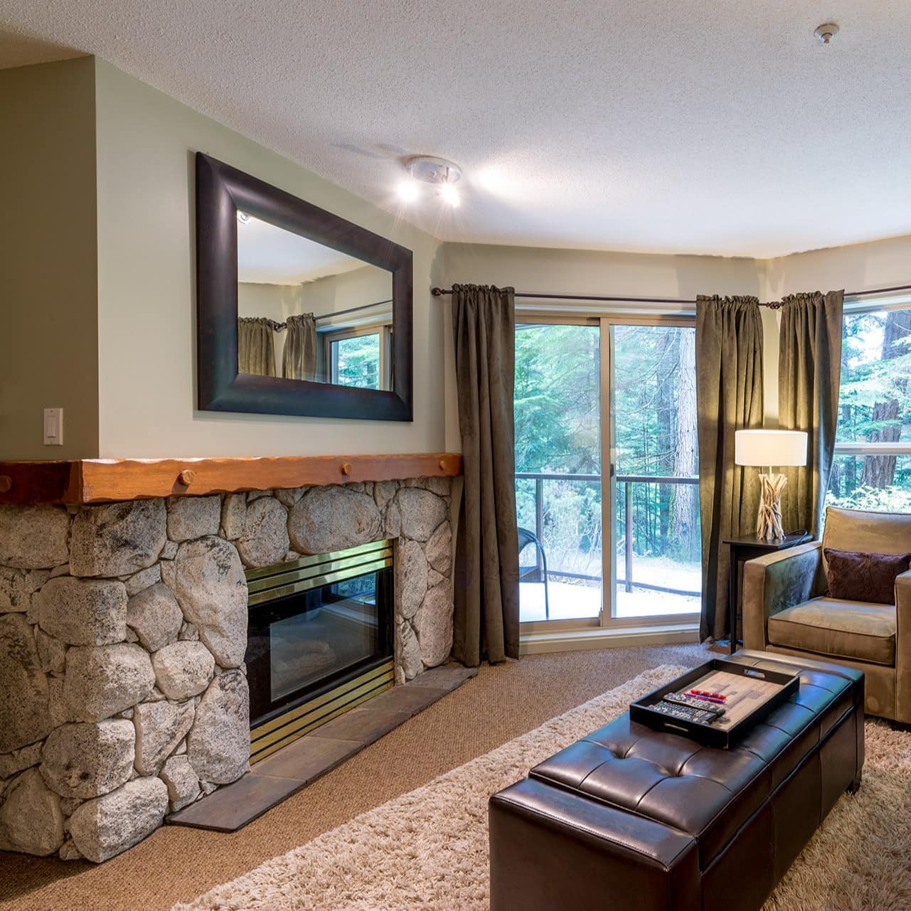 Spacious living room with comfortable furniture, stone fireplace, and 2 large windows looking into the forest