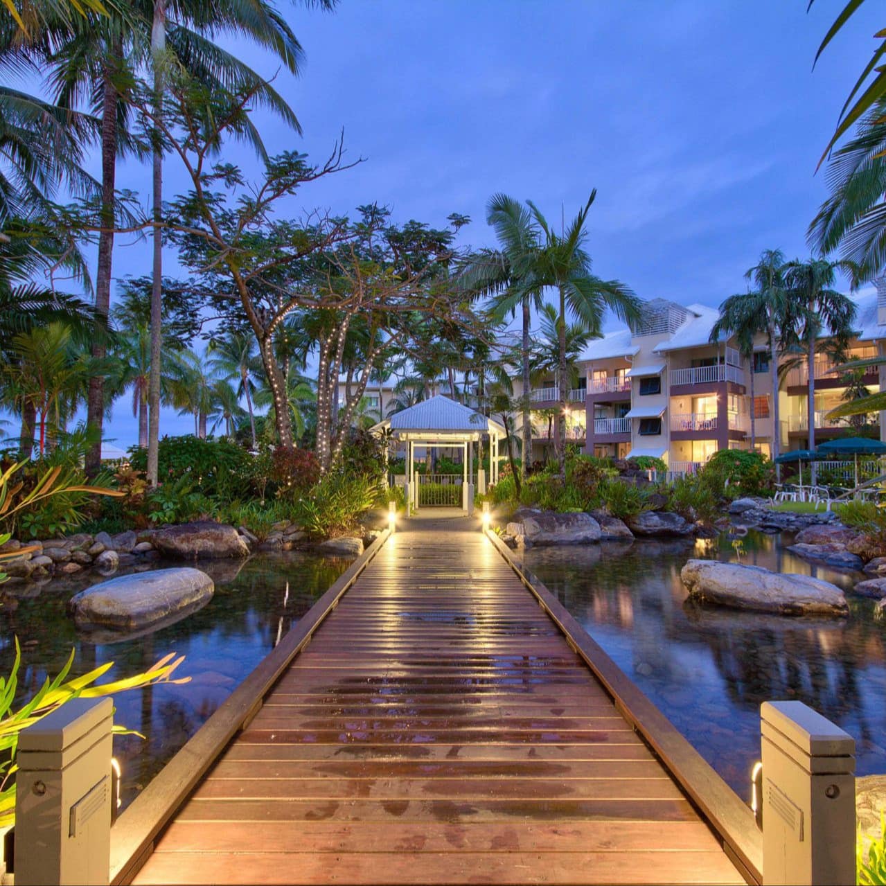 Apartment complex of 2-bedroom beach accommodation at sunset with boardwalk over pond and lighted palm trees